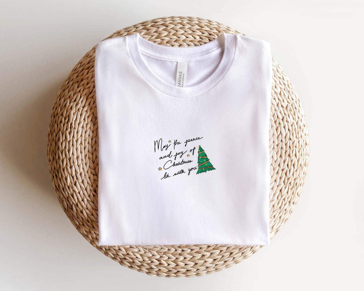 A white t-shirt with a Christmas tree design and text printed on it, placed on a woven basket.