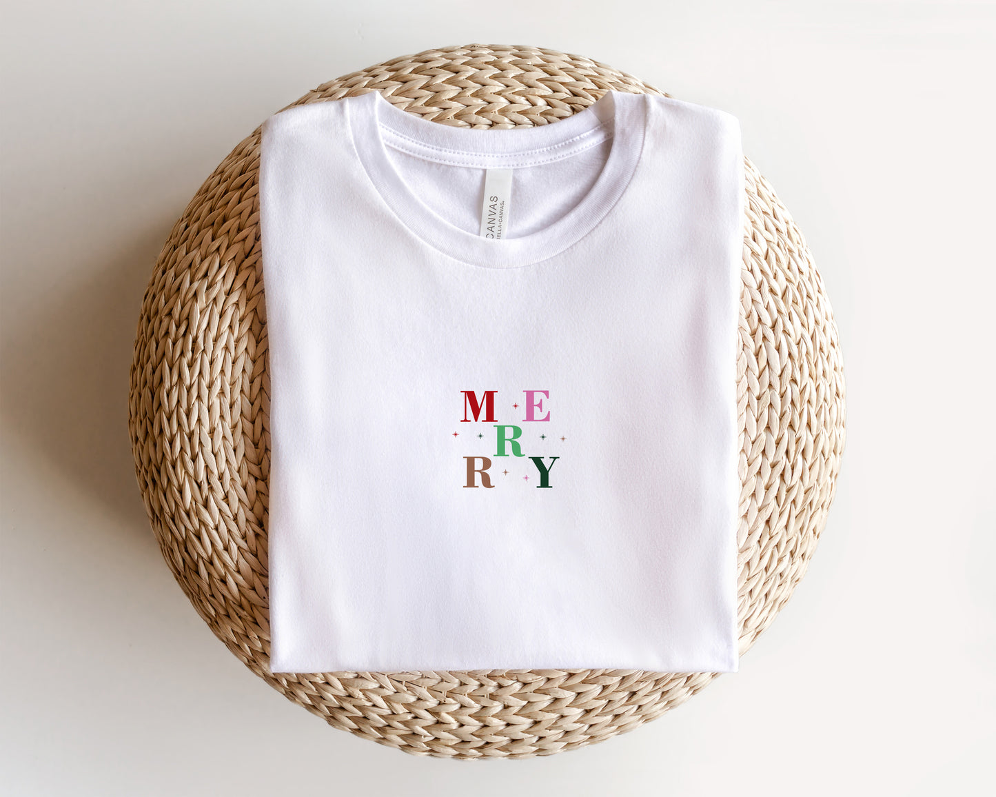 A white t-shirt with the word "MERRY" printed on it, laid out on a woven basket or mat.