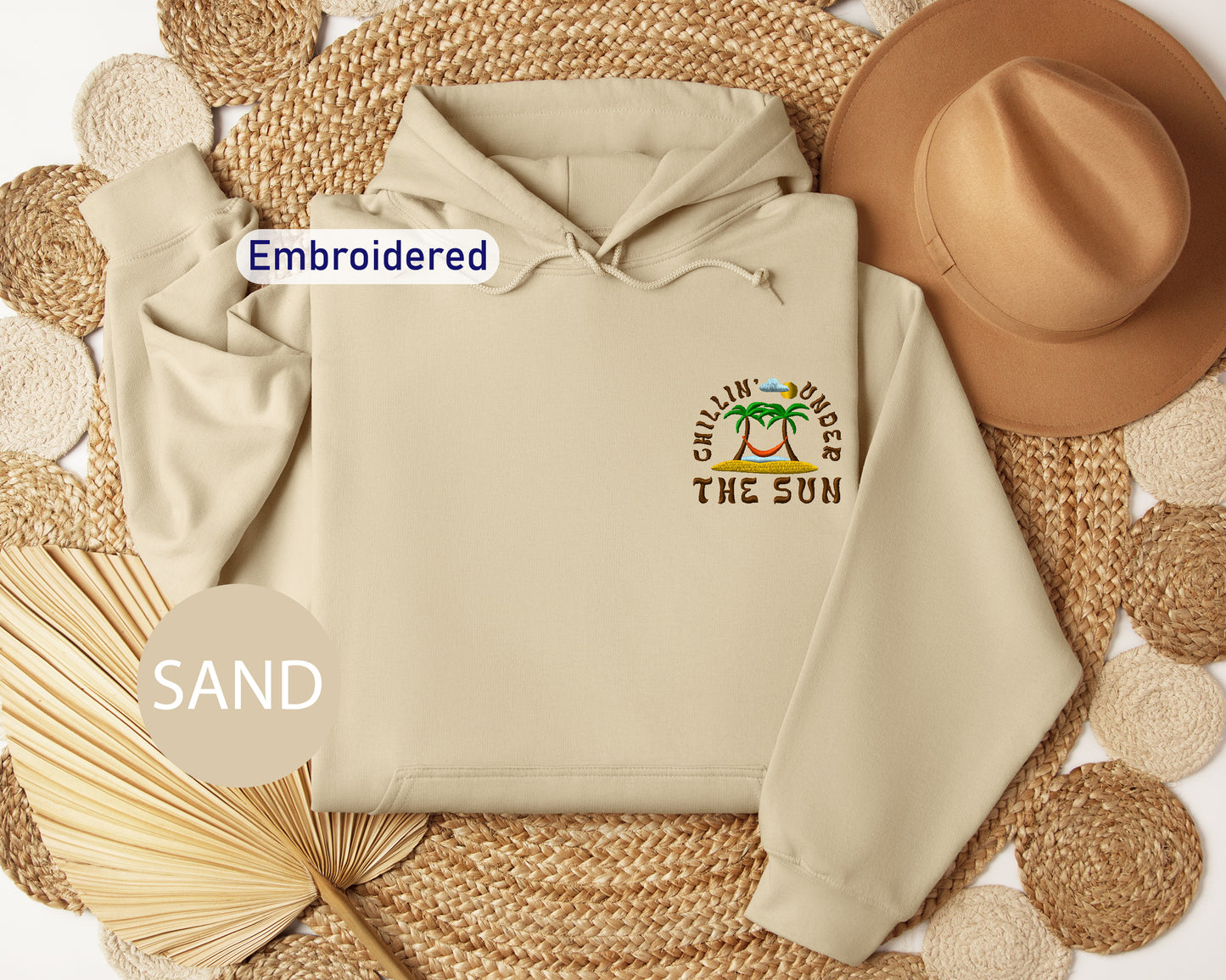 a beige hoodie with a tropical beach design, a brown hat, and a straw hat, all arranged on a woven mat.