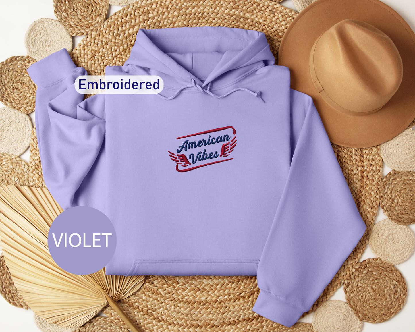 a purple hoodie with the text "American Vibes" embroidered on it, along with a brown hat and a straw hat placed on a woven mat.