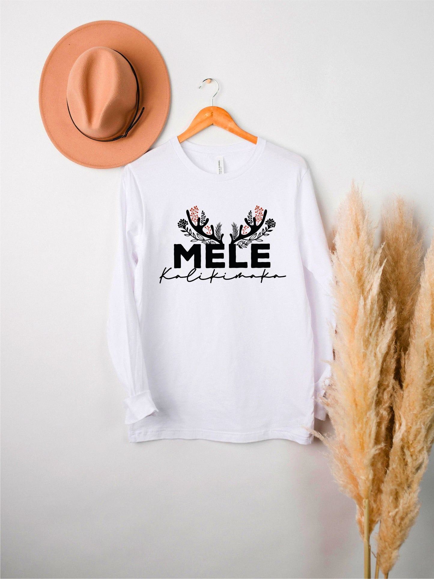 A white long-sleeved t-shirt with the text "MELE KOLLEK" printed on it, along with a brown hat and some potted plants in the background.