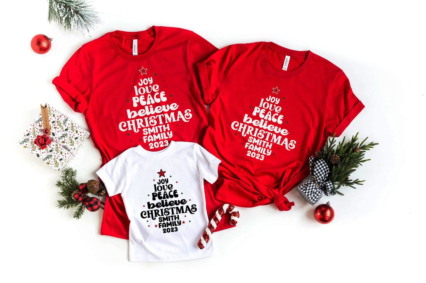 a collection of red t-shirts with Christmas-themed text, along with some Christmas decorations such as pine branches, ornaments, and a small white t-shirt.