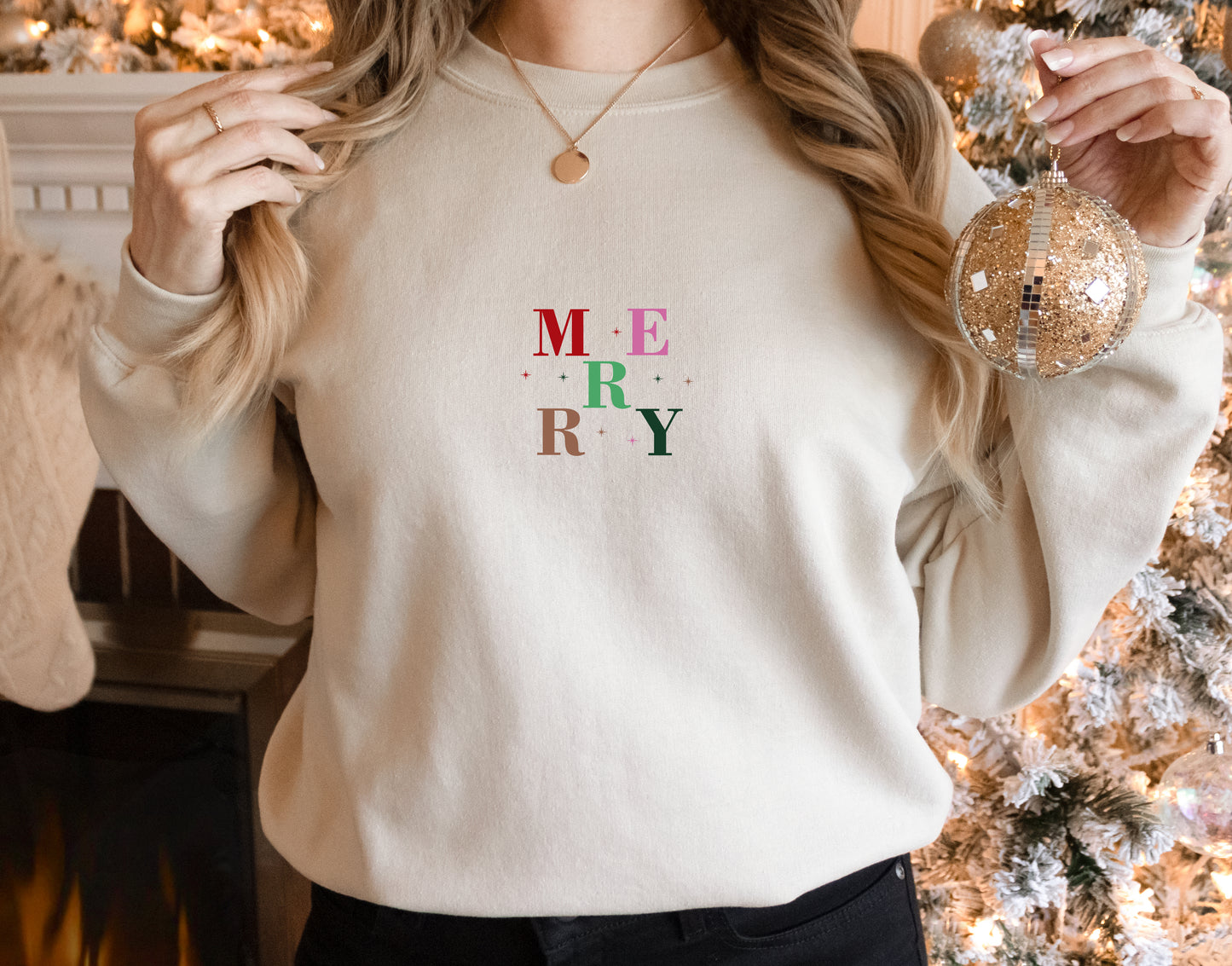 A woman wearing a white sweater with the word "MERRY" printed on it, holding a Christmas ornament in her right hand.