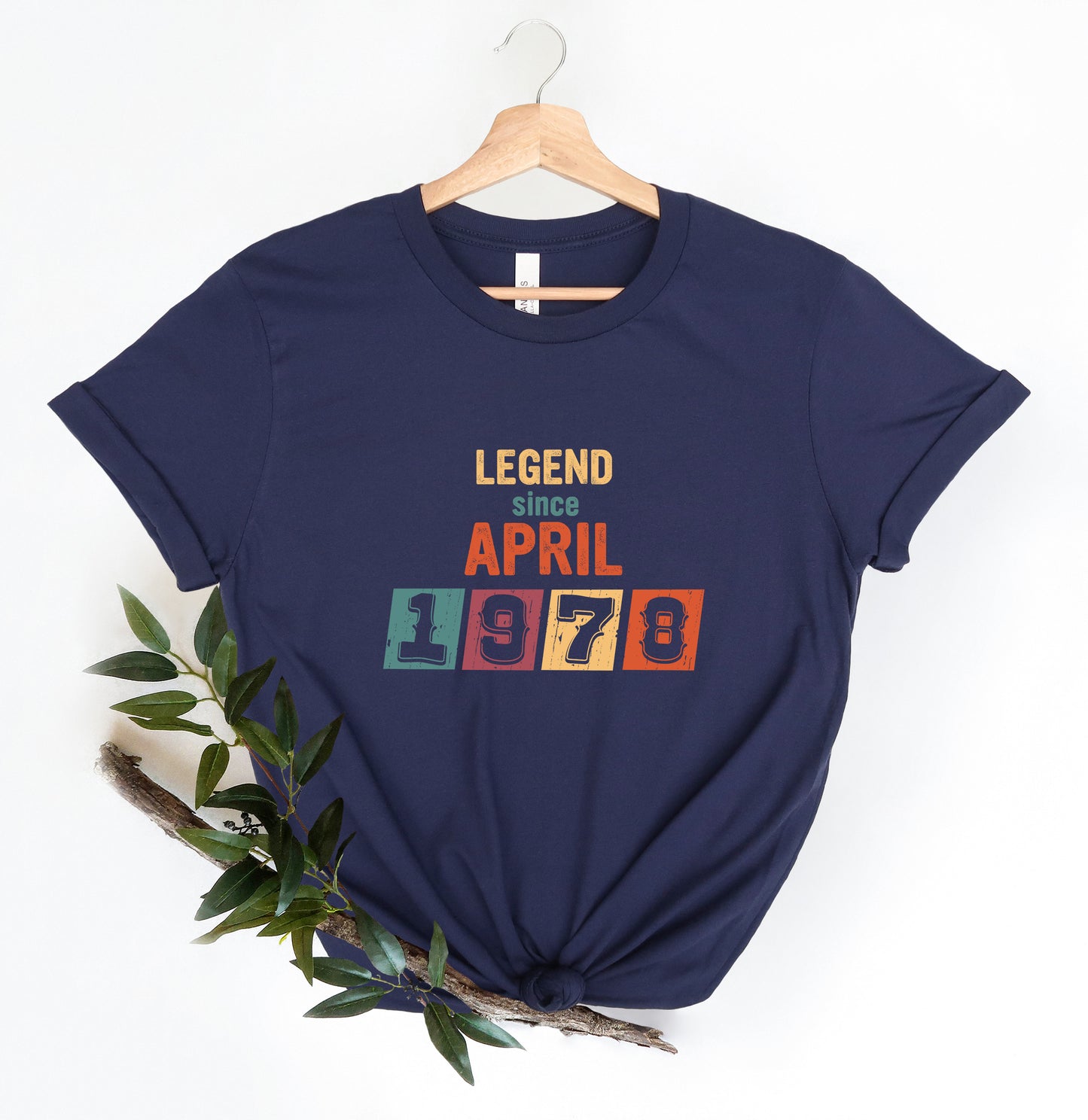 A navy blue t-shirt with the text "LEGEND since APRIL 1978" printed on it, along with a colorful block design. The t-shirt is hanging on a wooden hanger, and there is a branch with green leaves next to it.