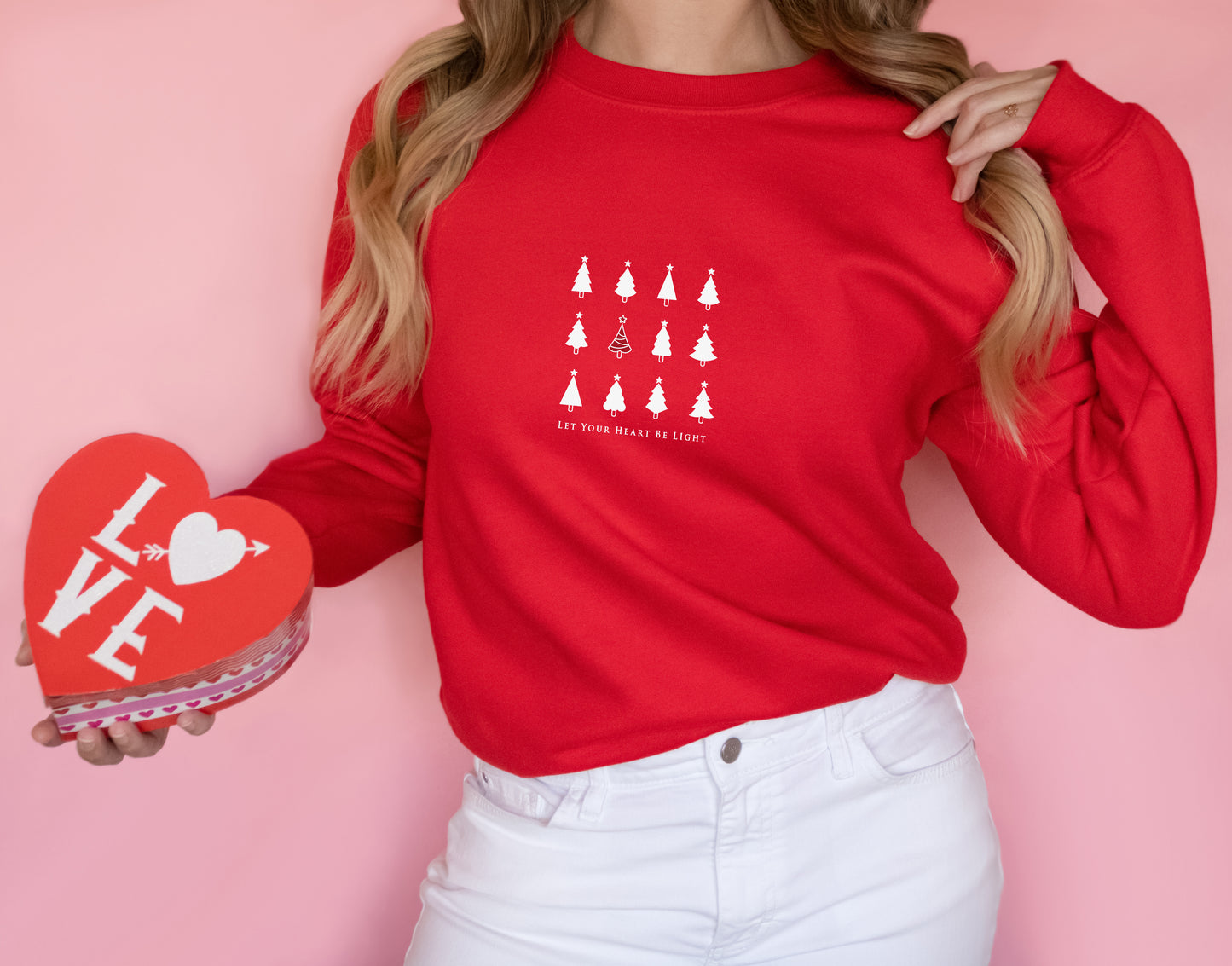 A woman wearing a red sweater with a white Christmas tree design and holding a red heart-shaped box with the word "LOVE" written on it.