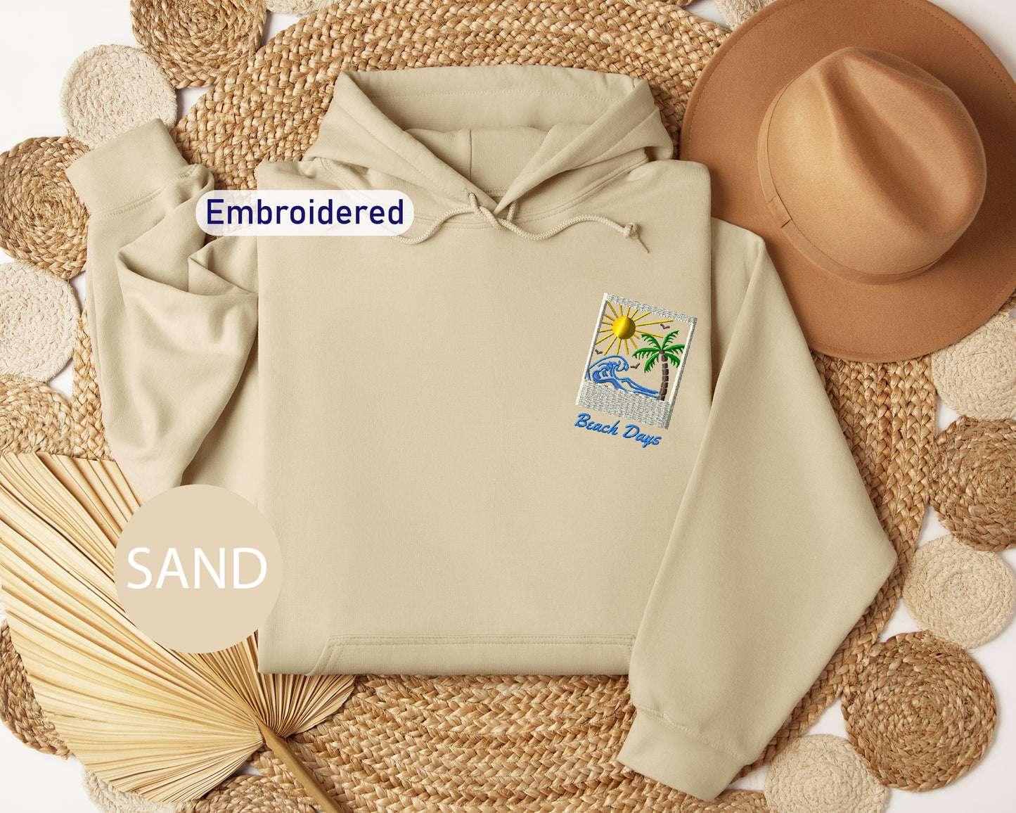 a beige hoodie with a beach-themed patch, a brown hat, and a fan, all arranged on a woven mat.