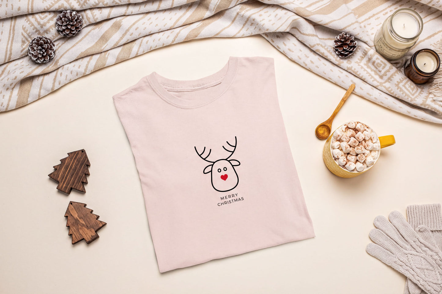 A pink t-shirt with a cartoon reindeer and the text "Merry Christmas" is displayed on a table, surrounded by holiday decorations such as pine cones, a candle, and a mug of hot chocolate.