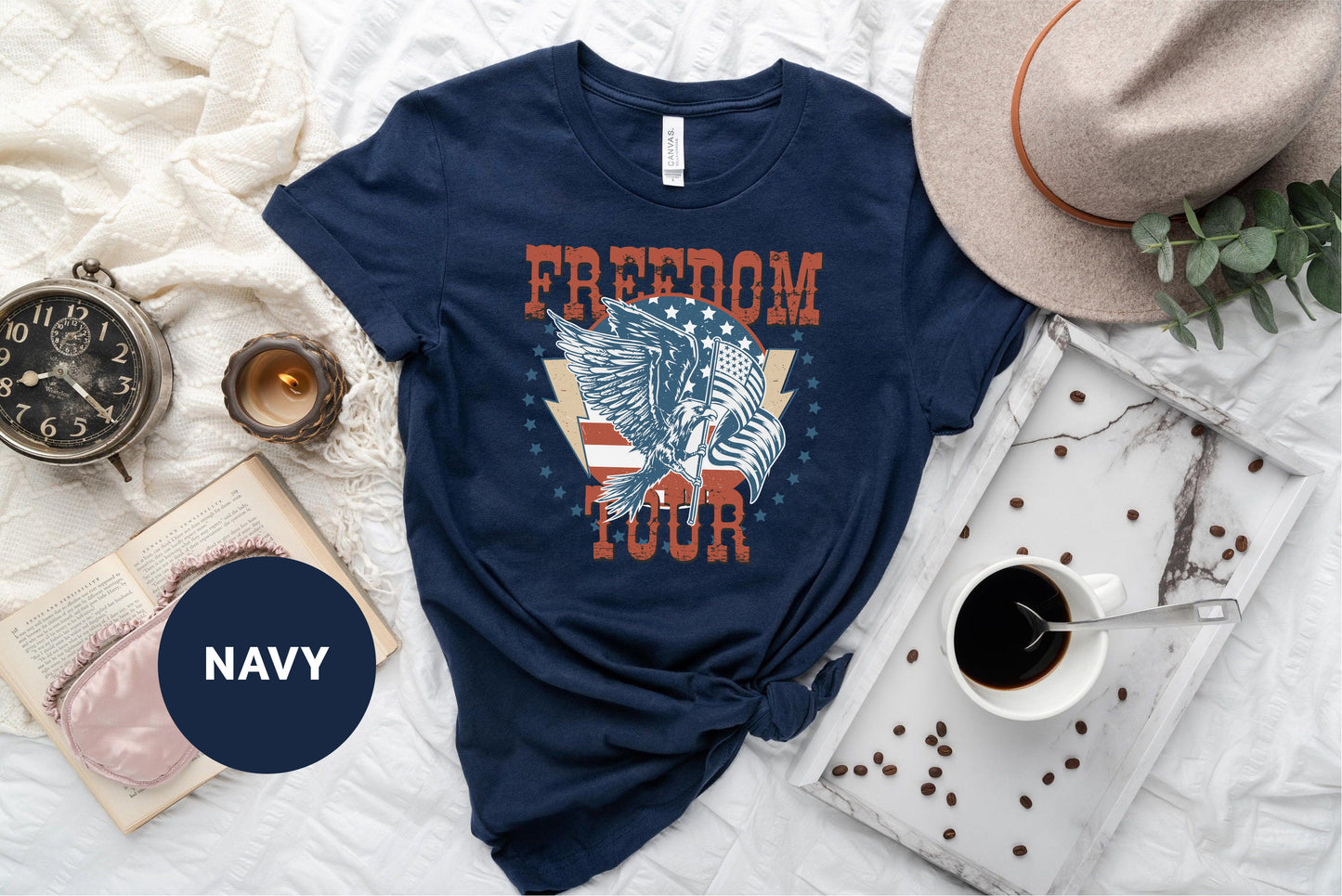 a navy blue t-shirt with a graphic design of a winged eagle and the word "FREEDOM TOUR" printed on it, along with a hat, a book, a cup of coffee, and some coffee beans.