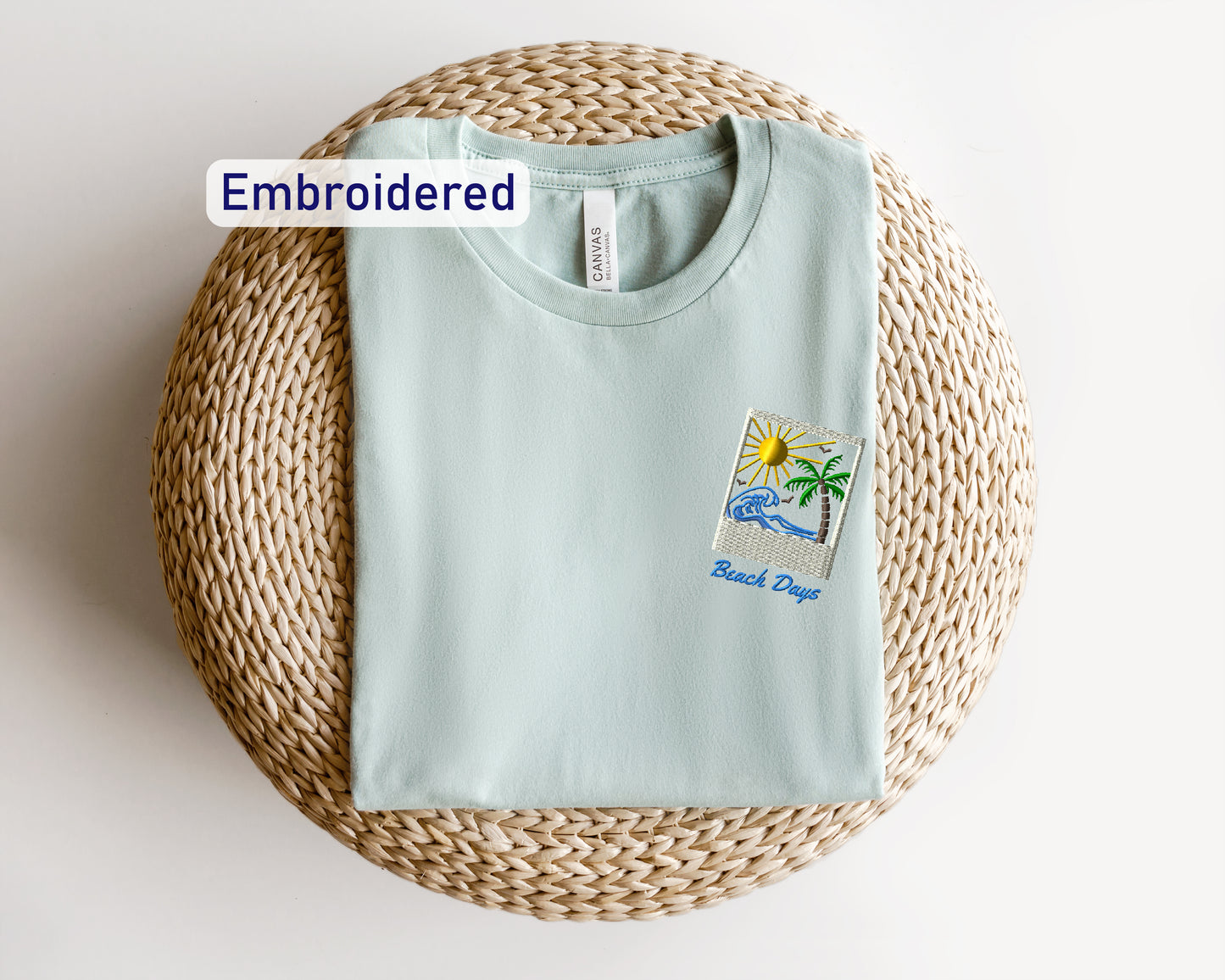 A light blue t-shirt with a beach-themed embroidered patch is displayed on a woven basket.