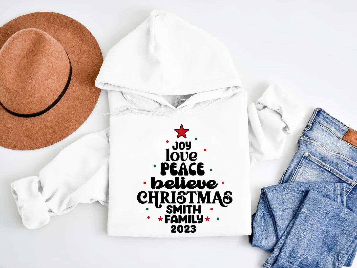 A white hoodie with a Christmas tree design and text, a brown hat, and a pair of blue jeans are arranged on a white background.