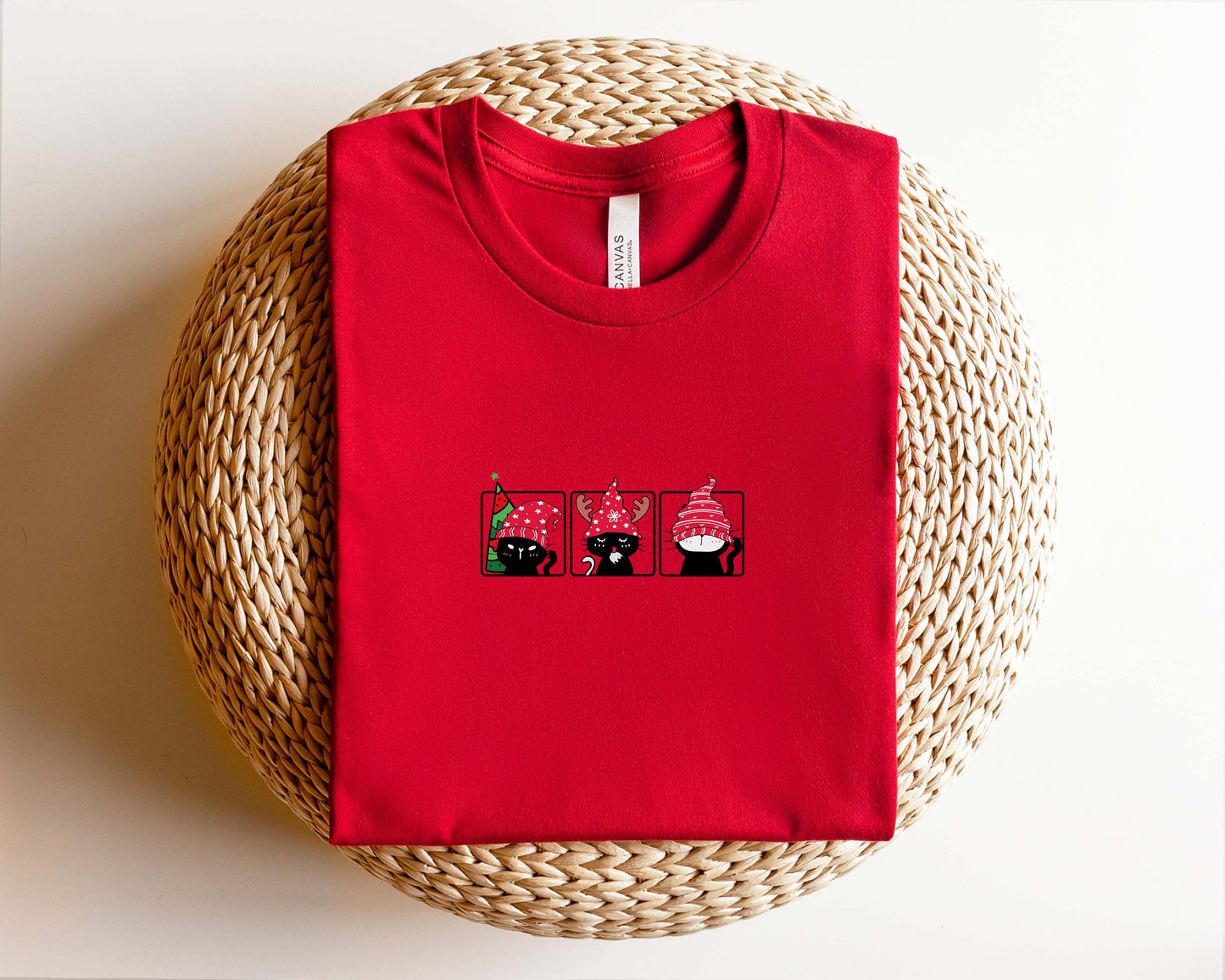 A red t-shirt with a graphic design of three cartoon cats wearing hats, displayed on a woven basket.