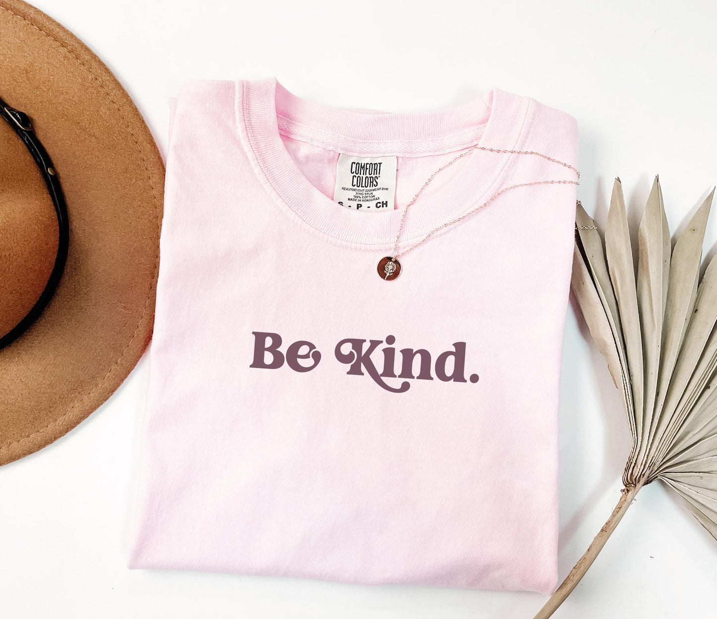 a pink shirt that says be kind next to a hat