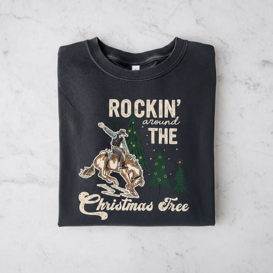 A black t-shirt with a graphic design depicting a cowboy riding a bucking bronco, with Christmas trees and the phrase "ROCKIN' around THE CHRISTMAS Tree" printed on it.