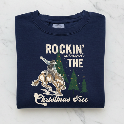 A navy blue sweatshirt with a graphic design depicting a cowboy riding a bucking bronco, with Christmas trees in the background. The text on the sweatshirt reads "ROCKIN' around THE CHRISTMAS Tree".