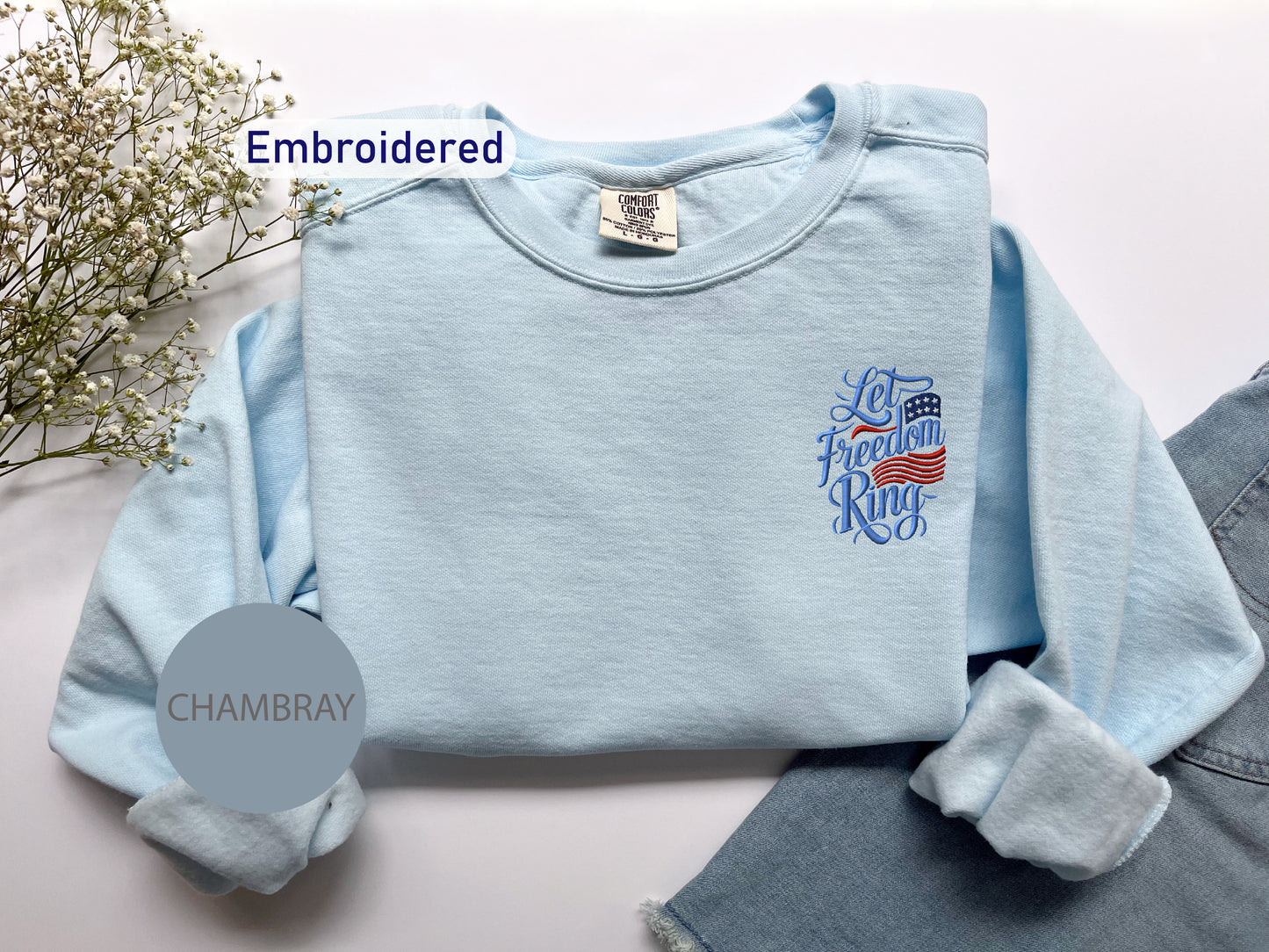 a light blue long-sleeved shirt with an embroidered design on the left chest area, placed on a white surface next to a pair of denim jeans.
