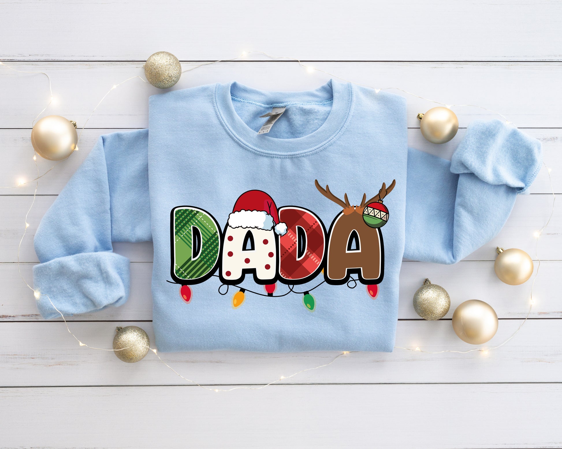 A light blue sweatshirt with the word "DADA" printed on it, surrounded by Christmas ornaments and lights on a white wooden surface.