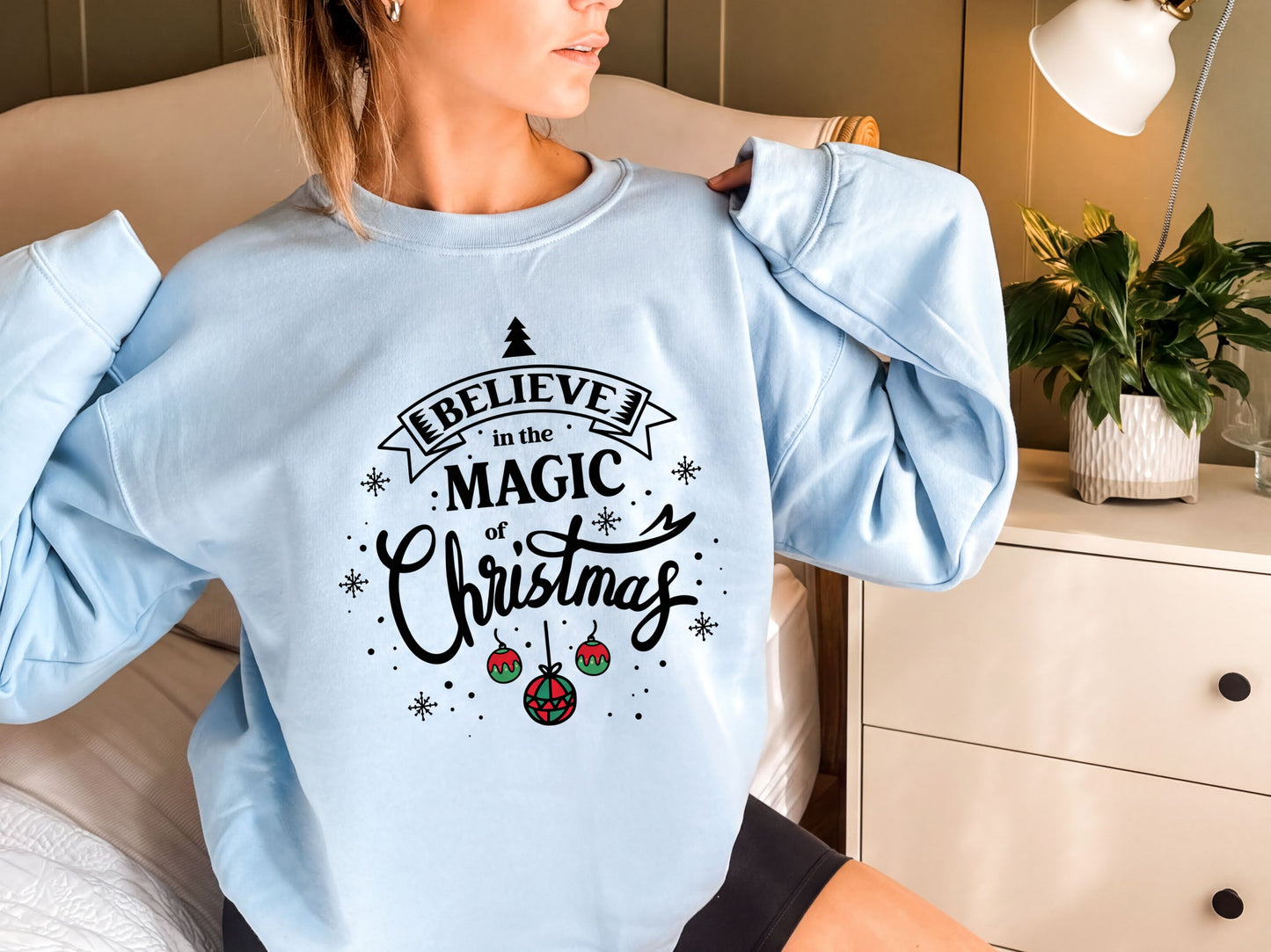 A woman wearing a light blue sweater with a printed design that says "Believe in the Magic of Christmas".