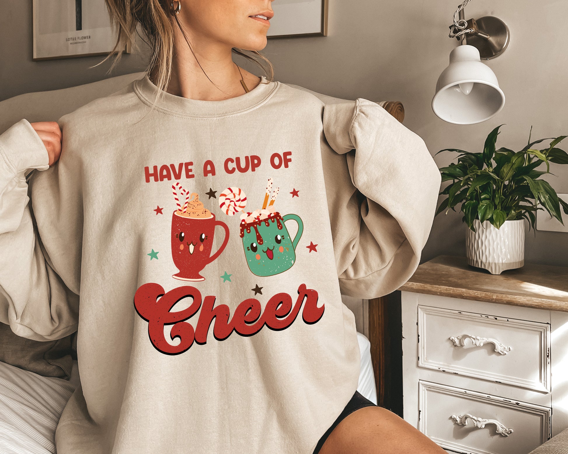 A woman is wearing a beige sweater with a graphic design that says "Have a cup of cheer" and features two colorful mugs with candy decorations.