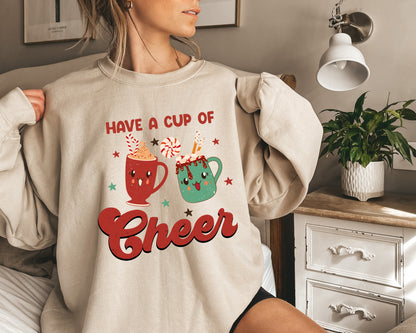 A woman is wearing a beige sweater with a graphic design that says "Have a cup of cheer" and features two colorful mugs with candy decorations.