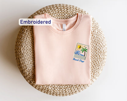 a pink t-shirt with a beach-themed embroidered patch on the front, placed on a woven basket or mat.