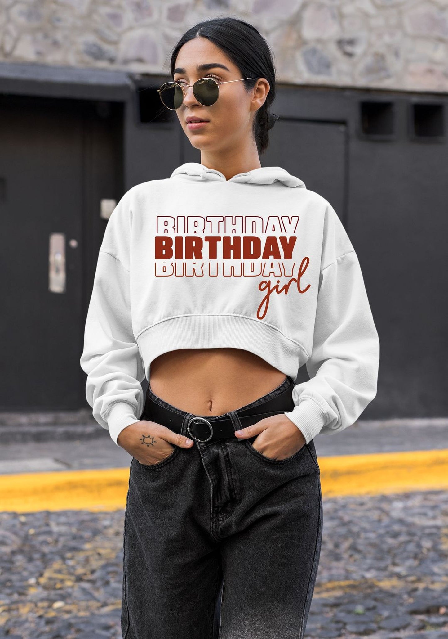 A woman wearing a white hoodie with the text "BIRTHDAY BIRTHDAY GIRL" printed on it, standing in front of a building.