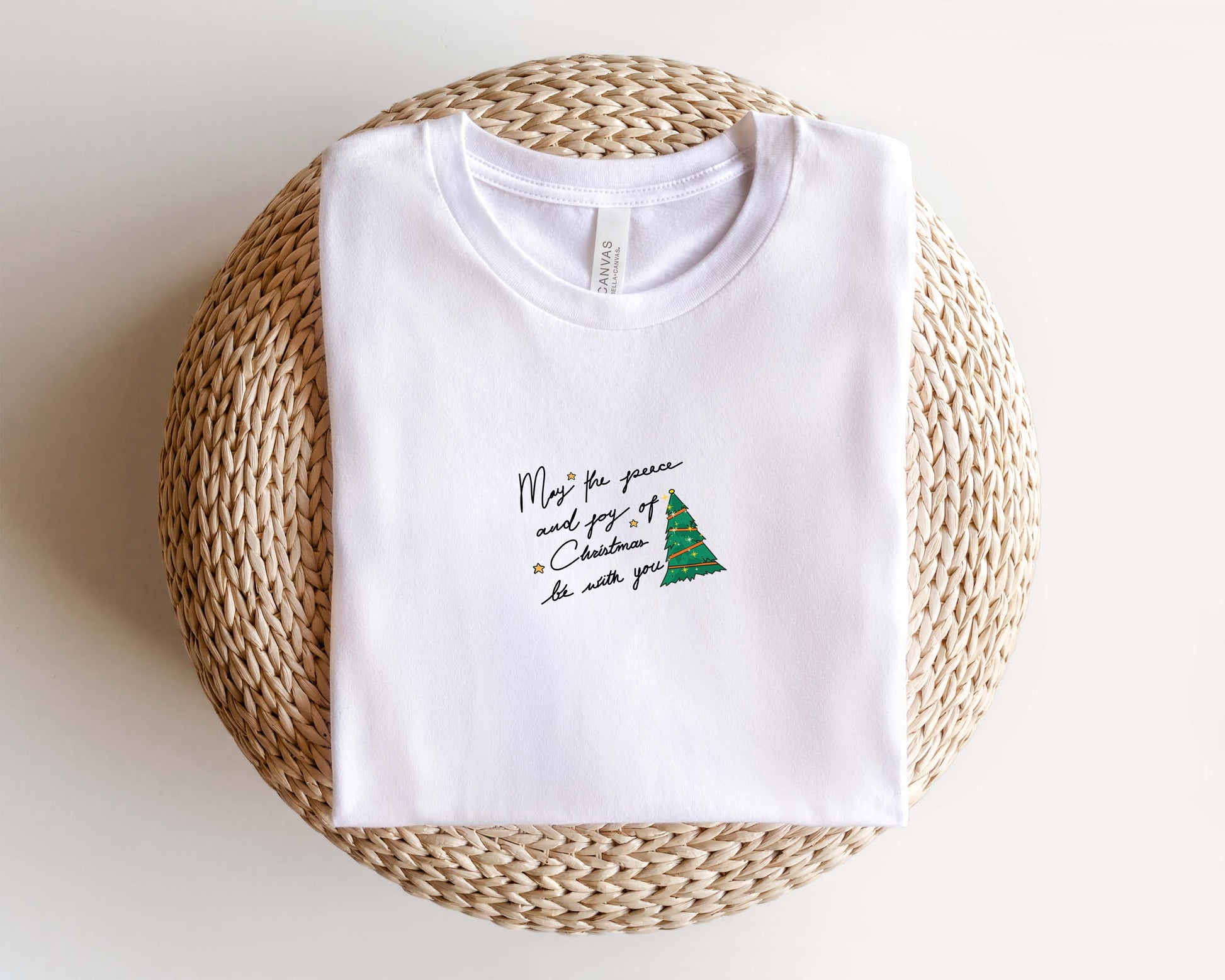A white t-shirt with a Christmas tree design and text printed on it, placed on a woven basket.