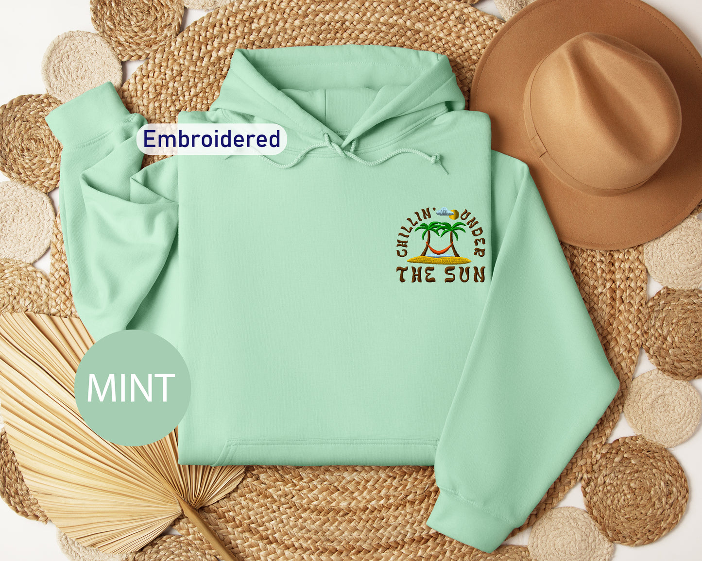 a mint green hoodie with a tropical beach-themed design, including palm trees and the phrase "Chillin' under the sun". The hoodie is embroidered with the text "Embroidered" and "MINT" is written in a circular shape at the bottom left corner.