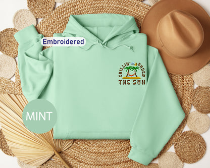 a mint green hoodie with a tropical beach-themed design, including palm trees and the phrase "Chillin' under the sun". The hoodie is embroidered with the text "Embroidered" and "MINT" is written in a circular shape at the bottom left corner.