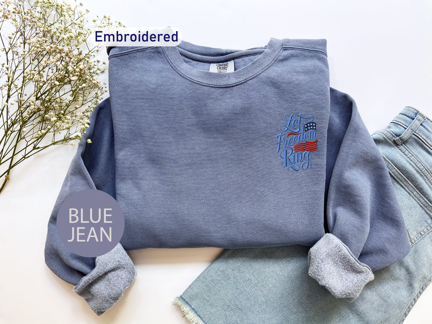 a blue sweatshirt with an embroidered design, a pair of blue jeans, and some flowers on a white background.