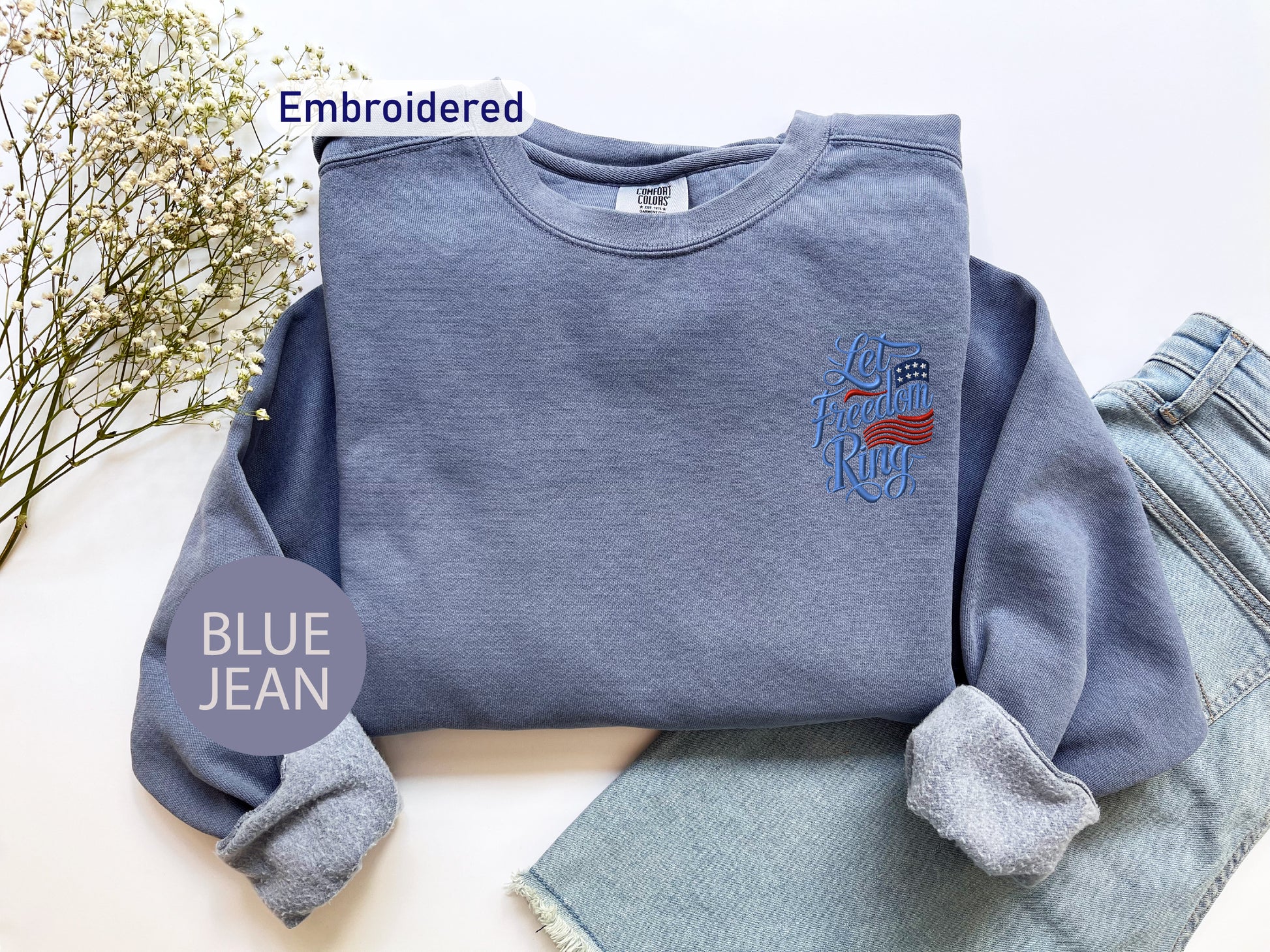 a blue sweatshirt with an embroidered design, a pair of blue jeans, and some flowers on a white background.