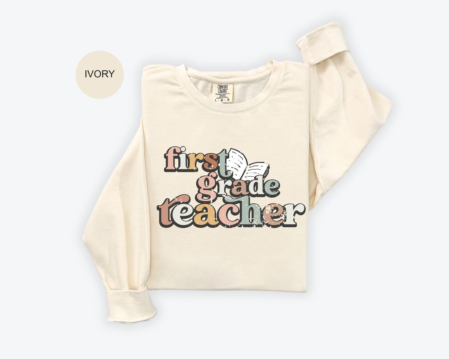 A beige long-sleeved t-shirt with a colorful graphic design that says "first grade teacher" printed on it.