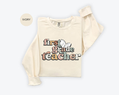 A beige long-sleeved t-shirt with a colorful graphic design that says "first grade teacher" printed on it.