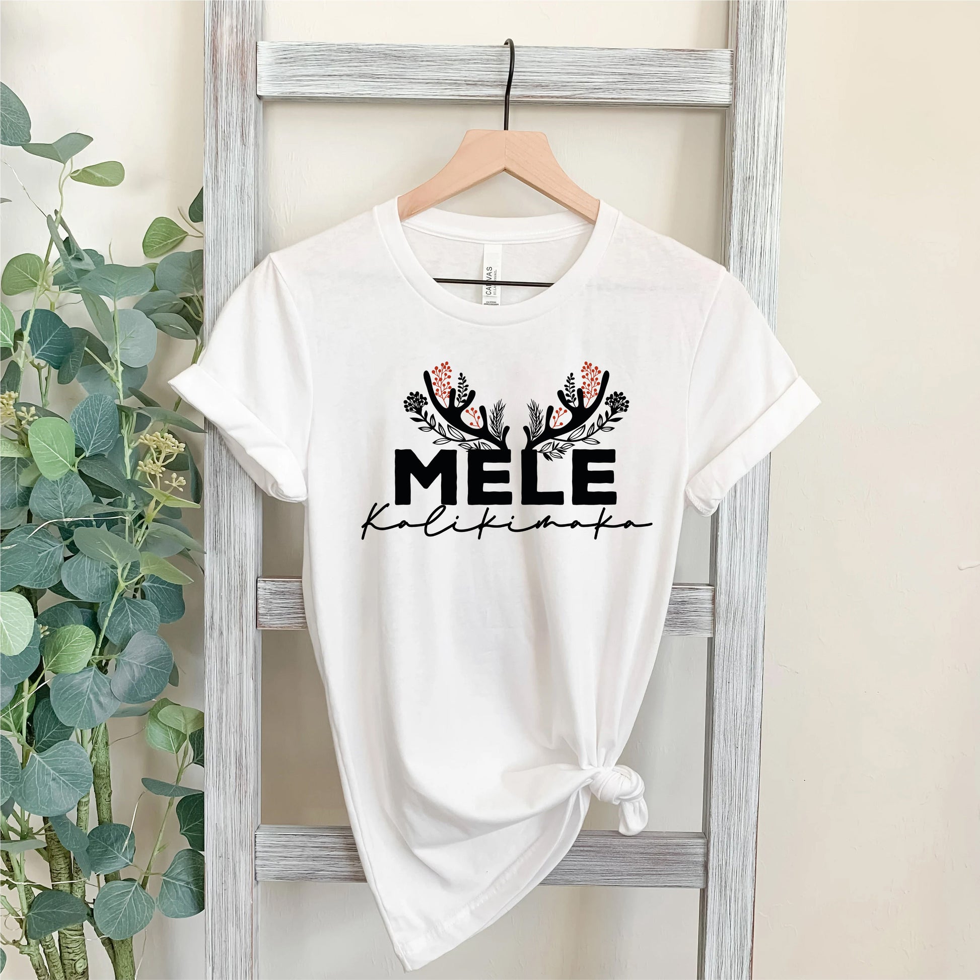 A white t-shirt with the text "MELE KILIKINAKO" printed on it, hanging on a wooden hanger.
