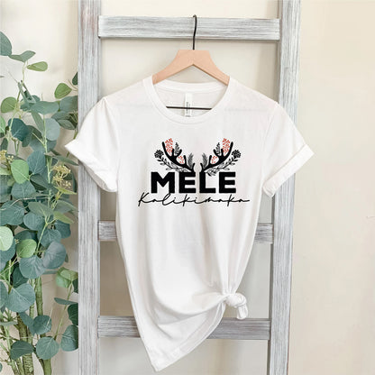 A white t-shirt with the text "MELE KILIKINAKO" printed on it, hanging on a wooden hanger.