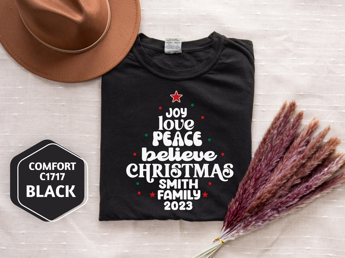 a black t-shirt with a Christmas tree design, a brown hat, and some pom poms, all laid out on a white surface.