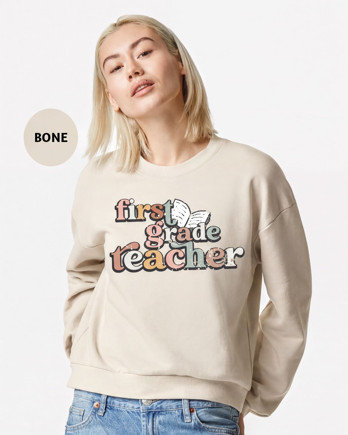 A woman wearing a beige sweater with the text "first grade teacher" printed on it.