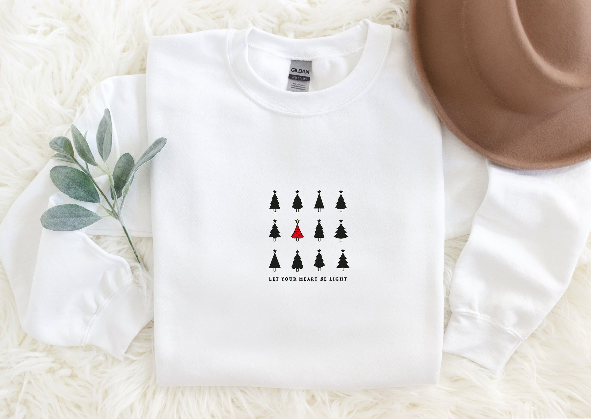 A white long-sleeved shirt with a graphic design of black Christmas trees and a red one, along with a brown hat, placed on a white fur-covered surface.