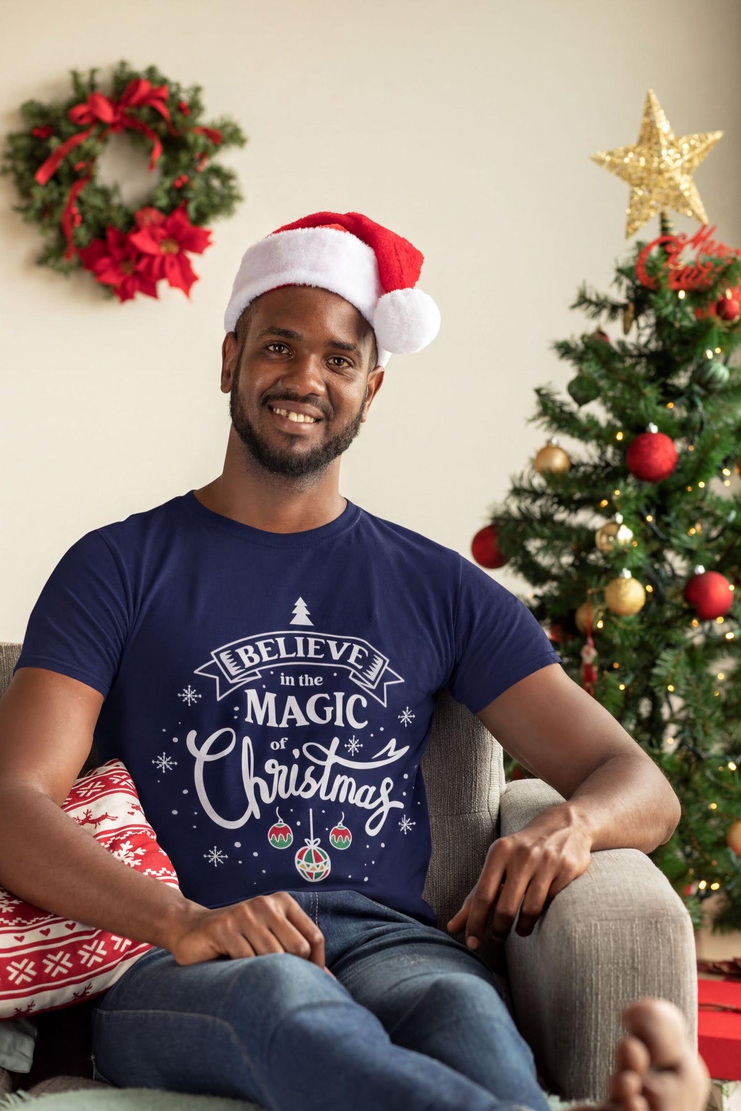 A smiling man wearing a Santa hat and a blue t-shirt with a Christmas-themed design is sitting in a chair in front of a Christmas tree.