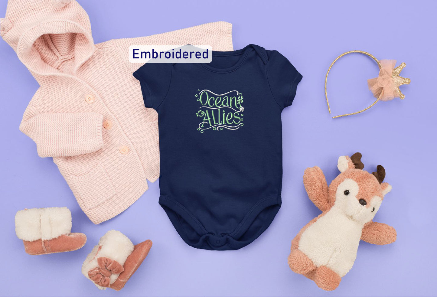 a collection of baby clothing and accessories, including a navy blue onesie with the text "Ocean Wives" embroidered on it, a pink hoodie, a gold headband, a pair of white booties, and a stuffed animal.