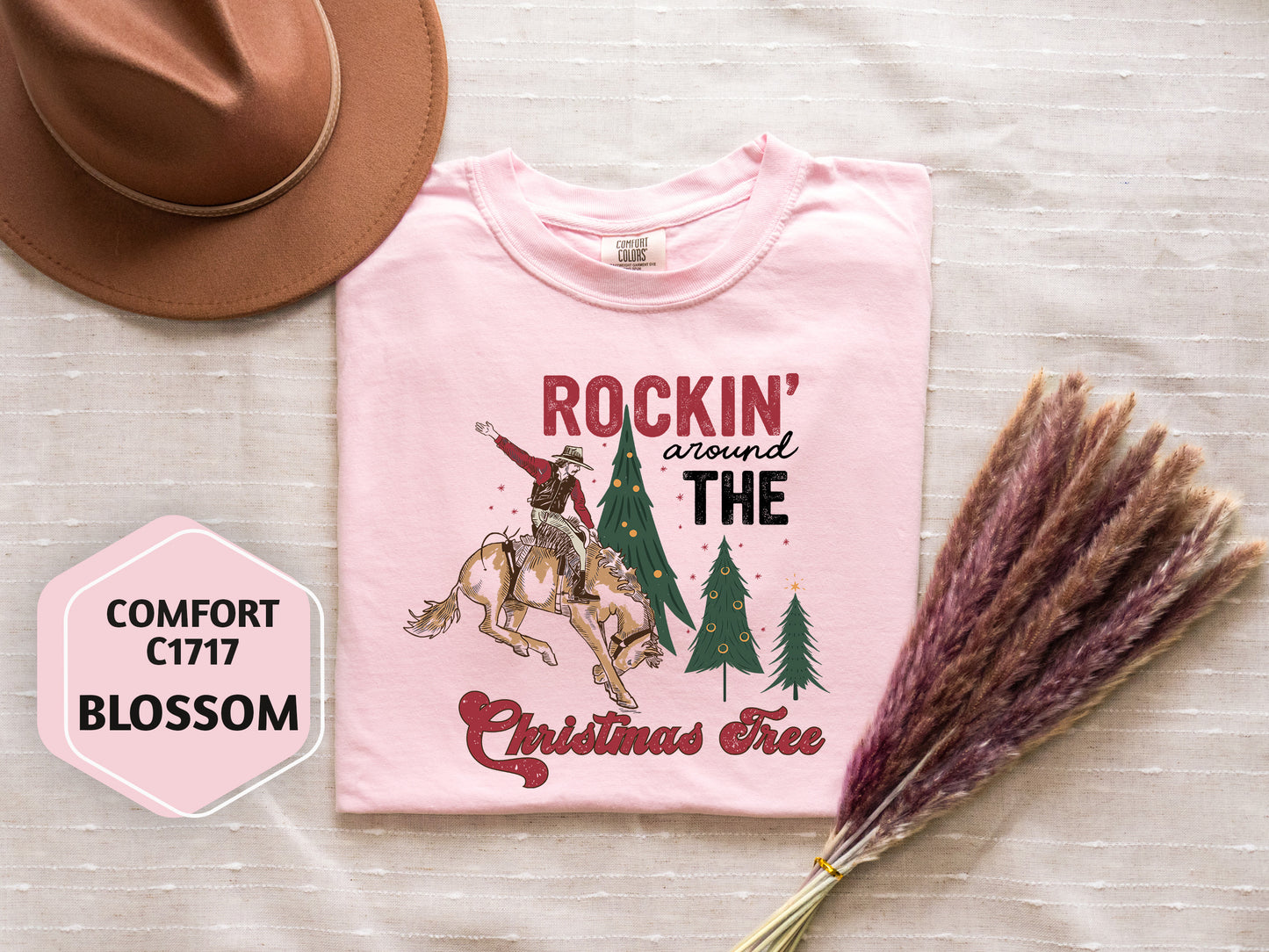 a pink t-shirt with a cowboy riding a bucking bronco, a cowboy hat, a pink flower, and a bunch of purple reeds, all arranged on a white background.