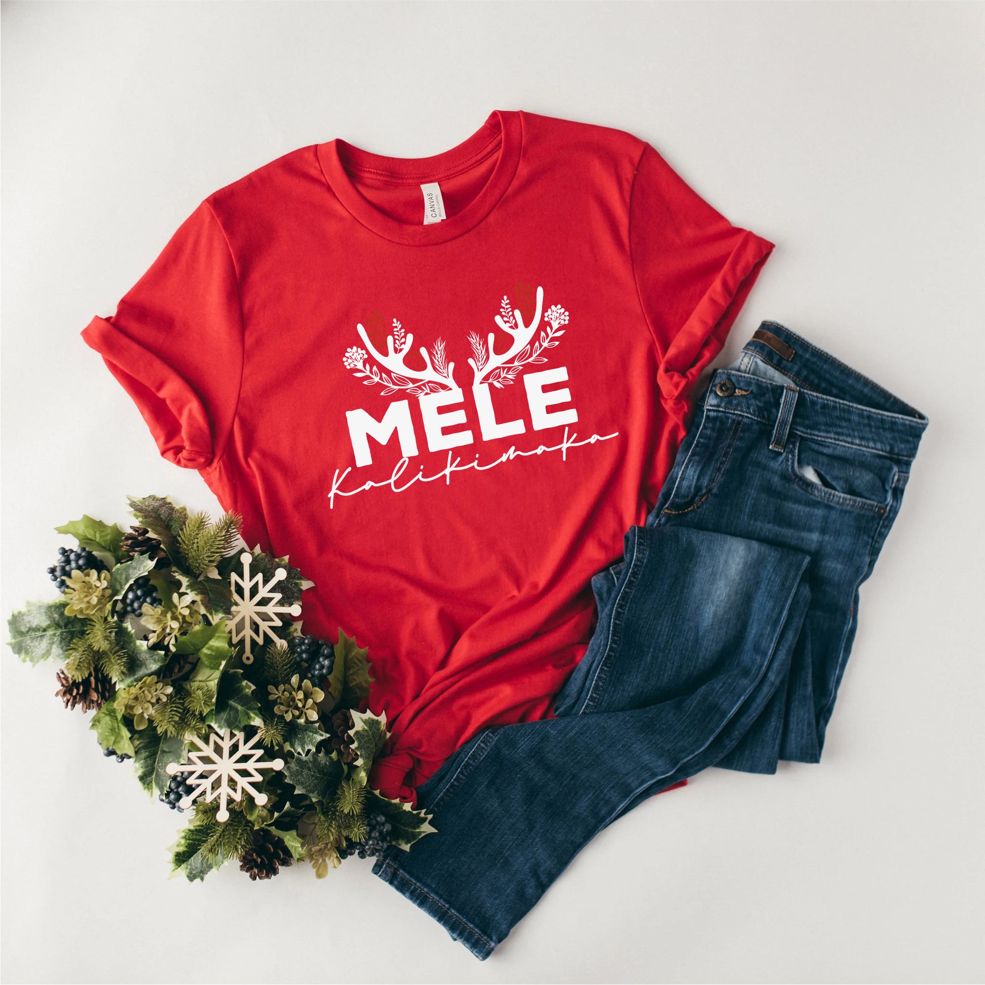 A red t-shirt with the text "MELE KOLIKINAS" printed on it, along with a pair of blue jeans and a wreath of pine cones.