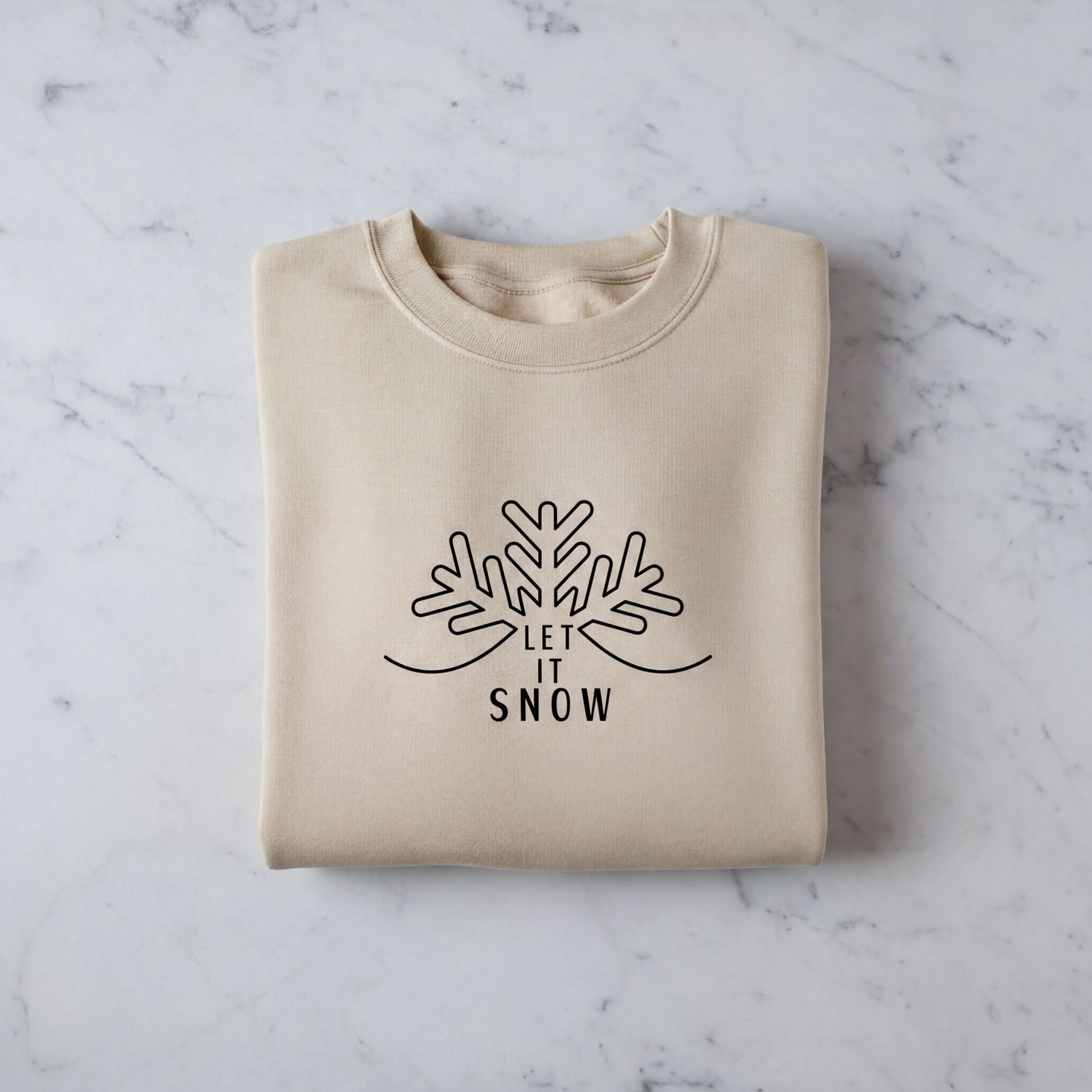 A beige sweater with a black snowflake design and the text "LET IT SNOW" printed on it, laid out on a marble surface.