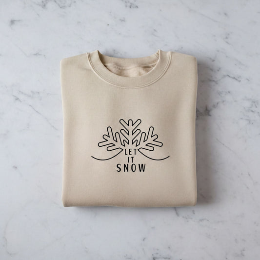 A beige sweater with a black snowflake design and the text "LET IT SNOW" printed on it, laid out on a marble surface.
