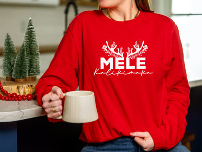 A person wearing a red sweater with the word "MELE" printed on it, holding a mug and standing in a kitchen setting with Christmas decorations visible in the background.