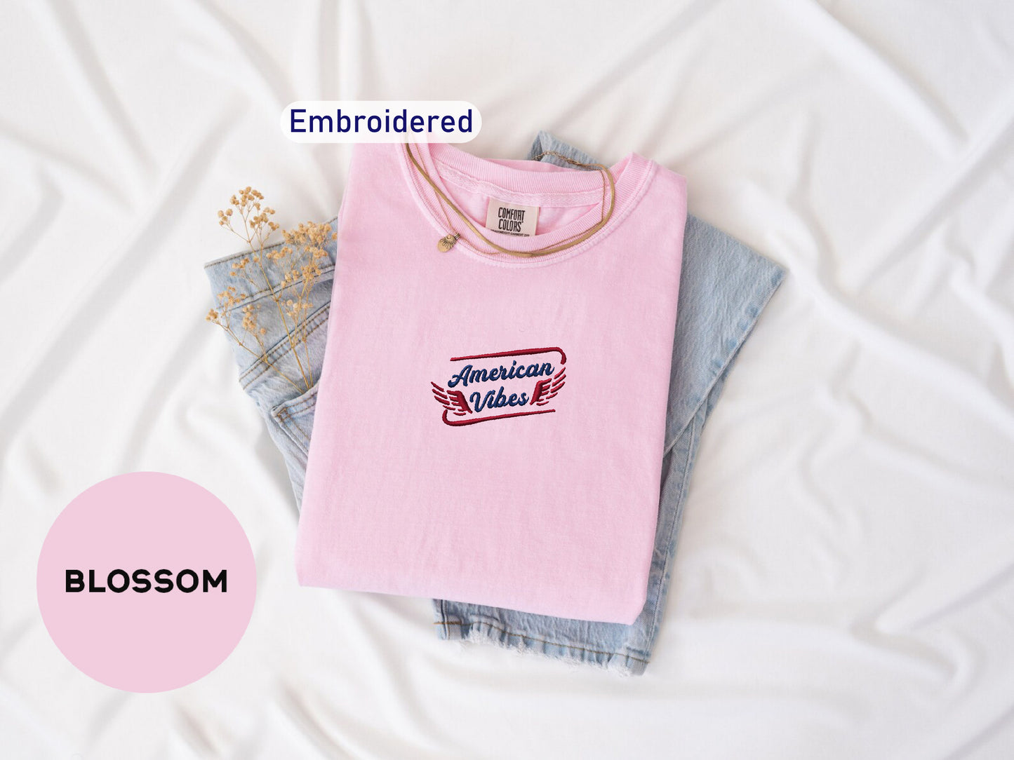 a pink t-shirt with the text "American Vibes" embroidered on it, along with a denim shirt and some flowers. The t-shirt is folded and placed on a white surface, with the text "Embroidered" and "BOMBSOM" visible.