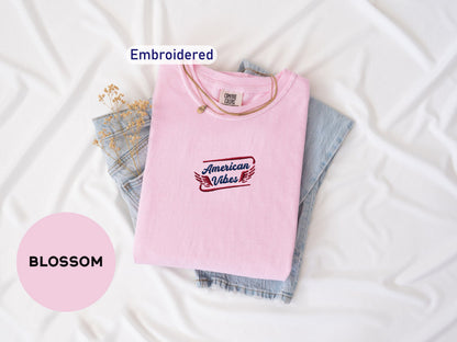 a pink t-shirt with the text "American Vibes" embroidered on it, along with a denim shirt and some flowers. The t-shirt is folded and placed on a white surface, with the text "Embroidered" and "BOMBSOM" visible.