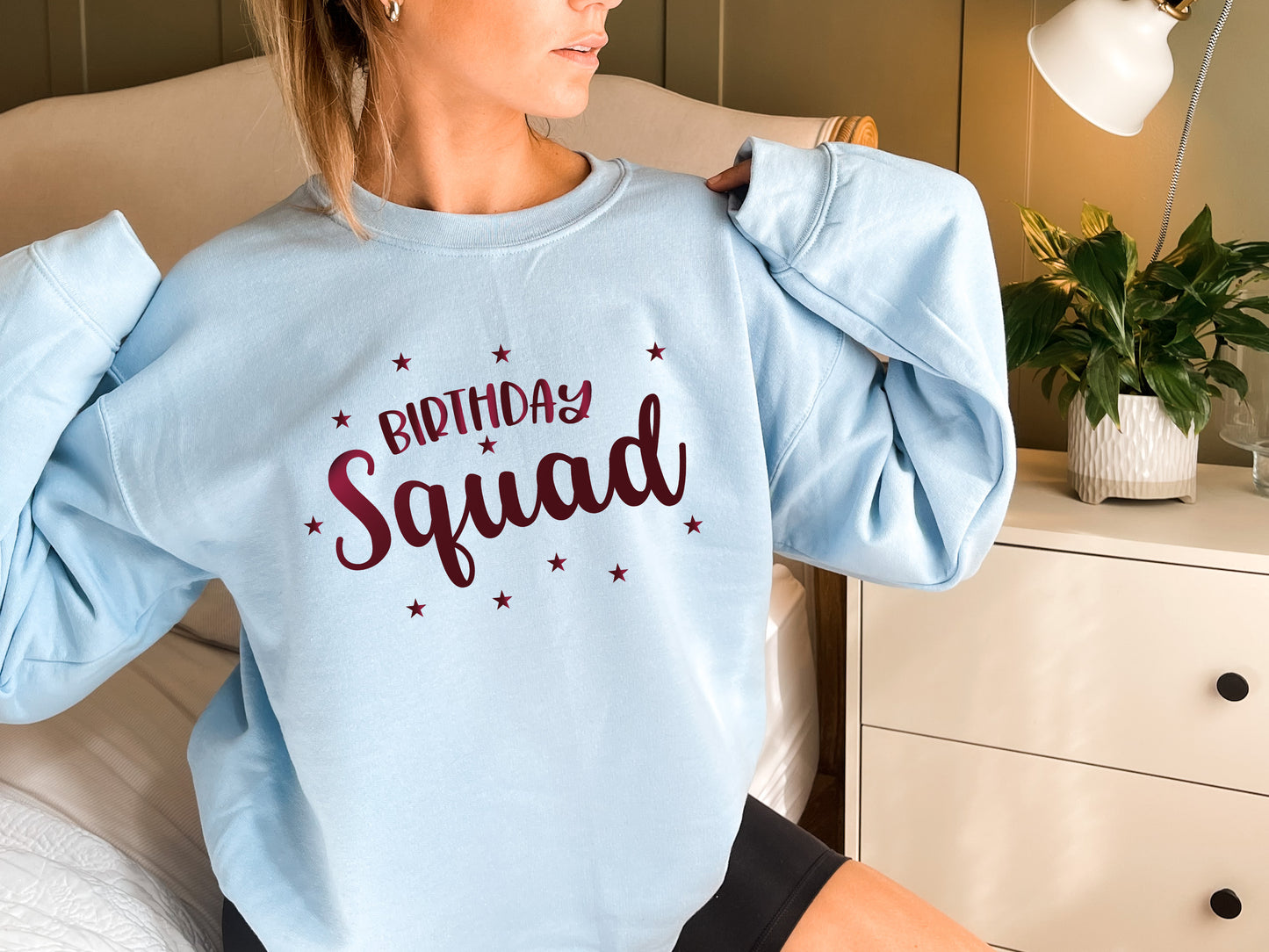 A woman wearing a light blue sweatshirt with the text "Birthday Squad" printed on it, posing in a bedroom setting with a potted plant and a dresser visible in the background.