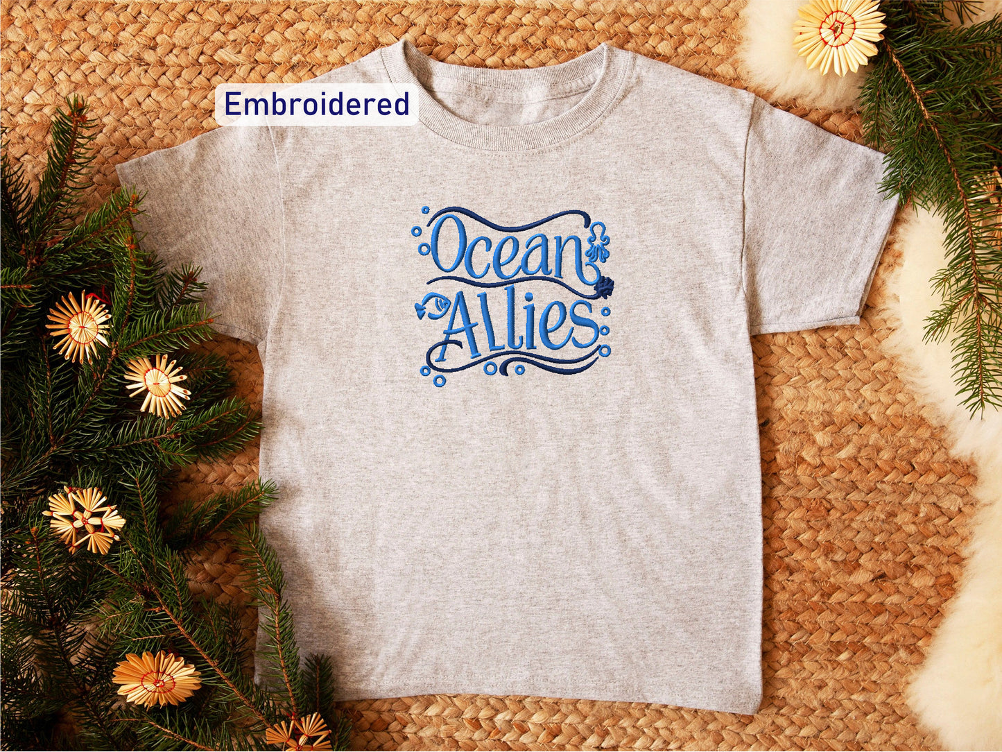 a gray t-shirt with the text "Ocean Allies" embroidered on it, placed on a woven surface surrounded by pine branches and flowers.