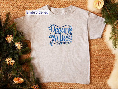 a gray t-shirt with the text "Ocean Allies" embroidered on it, placed on a woven surface surrounded by pine branches and flowers.