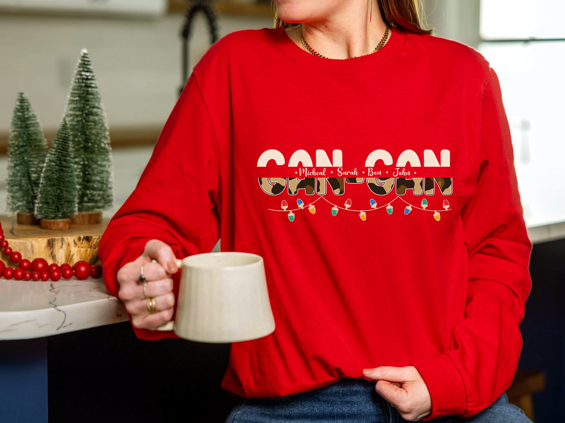 A person wearing a red sweater with the text "CAN CAN" is holding a white mug in a kitchen setting.